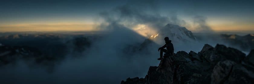 Una persona sentada en una cumbre rocosa sobre un mar de nubes, mirando hacia una montaña nevada más alta iluminada por el sol.
