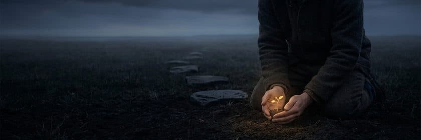 Una persona arrodillada en un campo oscuro protegiendo un brote luminoso entre las manos