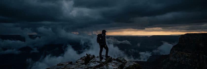 Un excursionista de pie en el borde de un acantilado mirando un valle cubierto de nubes de tormenta