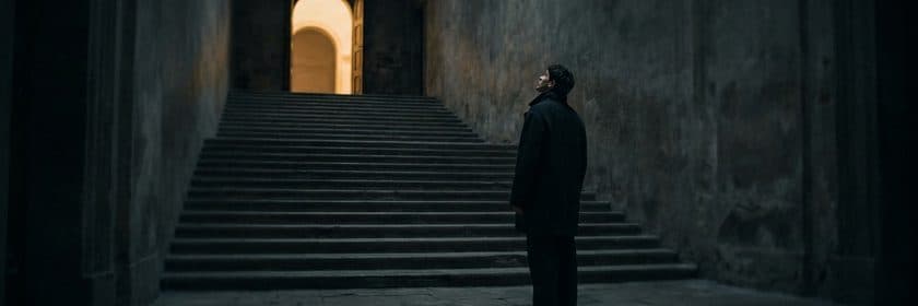 Una persona detenida al pie de una larga escalera de piedra, mirando hacia una puerta iluminada arriba