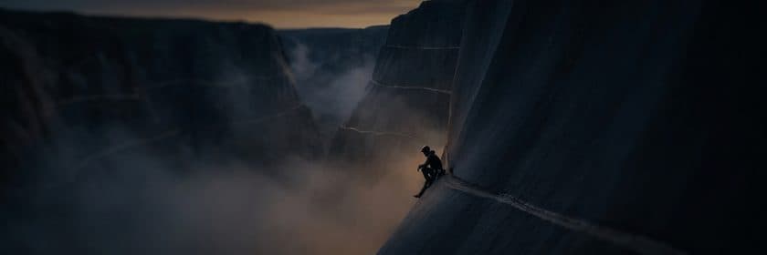 Un escalador sentado en una cornisa a media altura de un cañón profundo al anochecer, mirando la neblina que tapa el fondo.