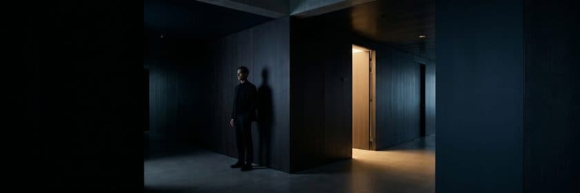 Una persona apoyada en la pared de un pasillo oscuro, junto a una puerta abierta con luz