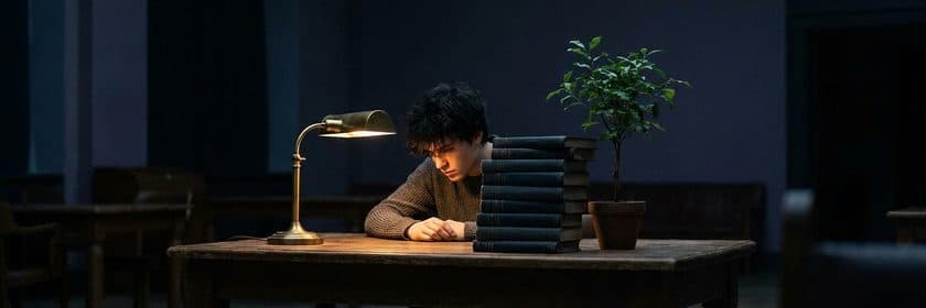 Un joven estudiando de noche en un escritorio junto a una pila de libros, una lámpara de bronce y una planta pequeña