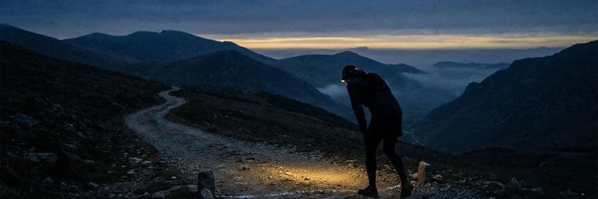 Una persona con linterna frontal recuperando el aliento al inicio de un sendero de montaña al amanecer
