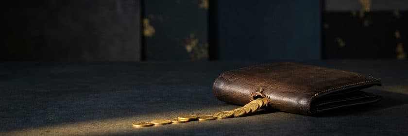 Una billetera de cuero rota por donde se escapa una fila de monedas doradas