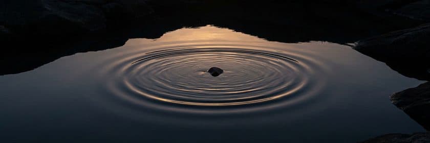 Una piedra que cae en agua quieta y abre anillos de ondas que reflejan una luz cálida