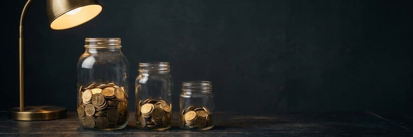 Tres frascos de vidrio con distintas cantidades de monedas bajo una lámpara de escritorio